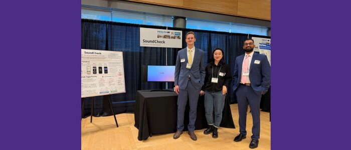 UW Master of Bioengineering students on the SoundCheck team standing at their table at a student competition. From left to right: Will Szymanski, Baolin Niu, Krishna Bathini