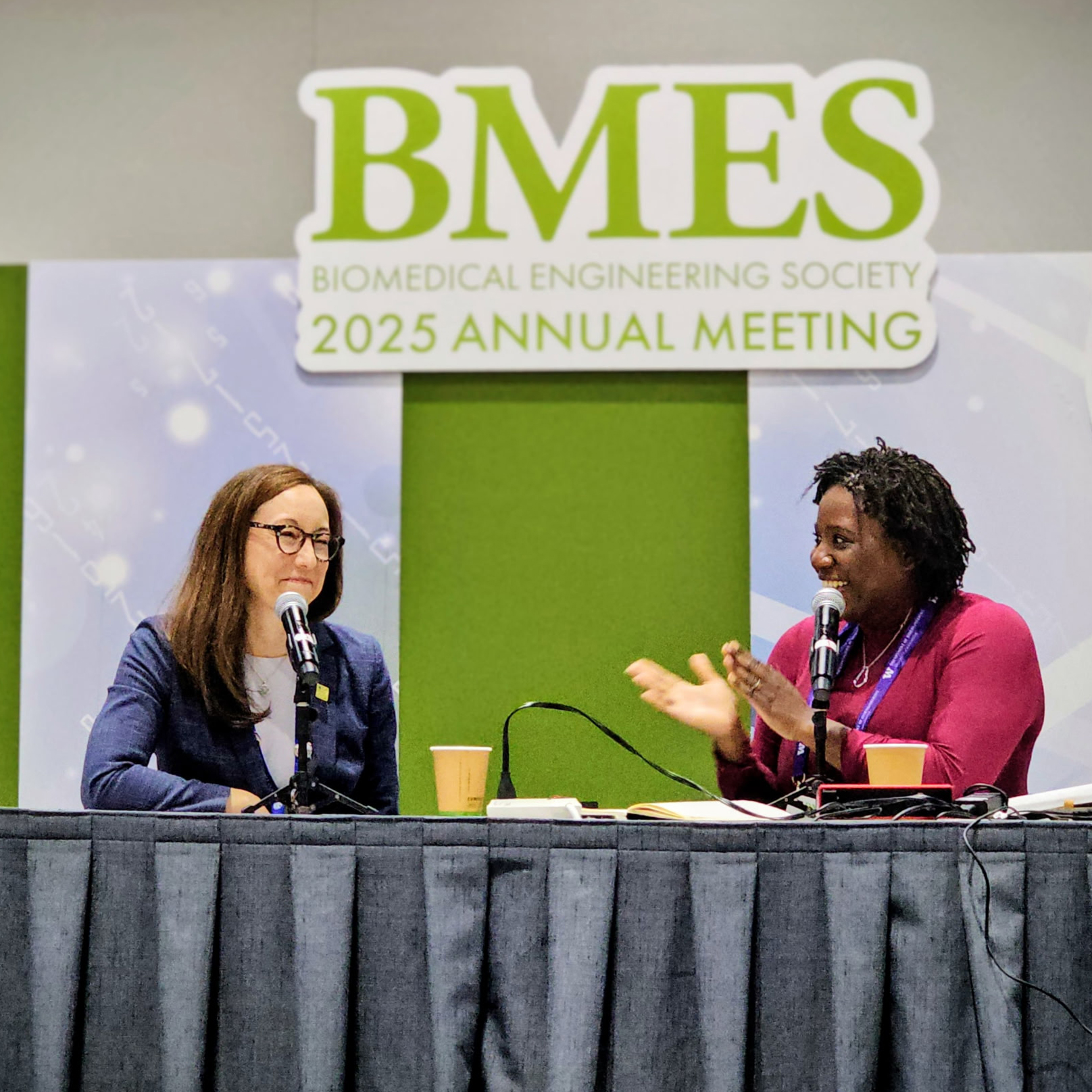Two women speak into microphones while sitting at a table. A sign behind them reads "BMES 2025 Annual Meeting."