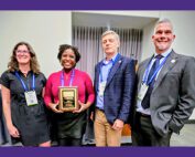 Four people smile for the camera while standing next to one another. A woman second from the left holds a rectangular plaque.
