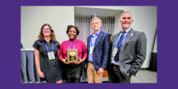 Four people smile for the camera while standing next to one another. A woman second from the left holds a rectangular plaque.