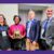 Four people smile for the camera while standing next to one another. A woman second from the left holds a rectangular plaque.